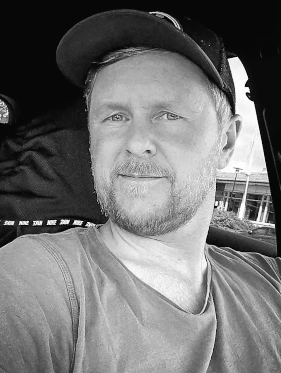 Person med svart caps og kort skjegg i en bil, med bygninger og parkeringsplass utenfor vinduet